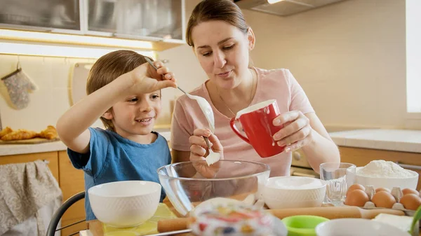小さな息子を持つ若い母親は大きなガラスボウルにパイやケーキを焼くための材料を注ぎます.両親、小さなシェフ、家族と一緒に時間を過ごす子供たち、家庭用キッチン. — ストック写真