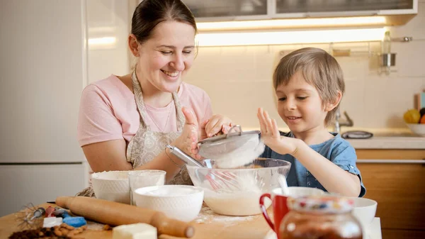 Маленький улыбающийся мальчик держит решето и помогает матери делать тесто для печенья. Дети готовят с родителями, маленький шеф-повар, семья, проводящая время вместе, домашняя кухня. — стоковое фото