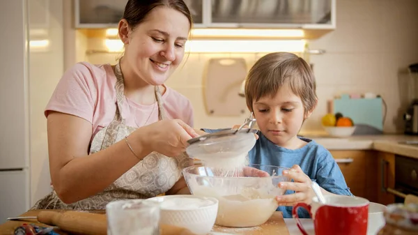 Giovane madre sorridente che cucina con il figlioletto e setaccia la farina con setaccio. Bambini che cucinano con i genitori, piccolo chef, famiglia che ha tempo insieme, cucina domestica. — Foto Stock