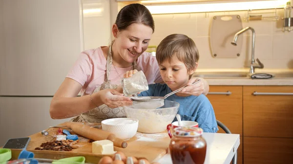 Νεαρή χαμογελαστή μητέρα μαθαίνει στο γιο της να κοσκινίζει αλεύρι με κόσκινο για να φτιάξει ζύμη μπισκότων. Παιδιά μαγείρεμα με τους γονείς, λίγο σεφ, οικογένεια έχοντας χρόνο μαζί, οικιακή κουζίνα. — Φωτογραφία Αρχείου