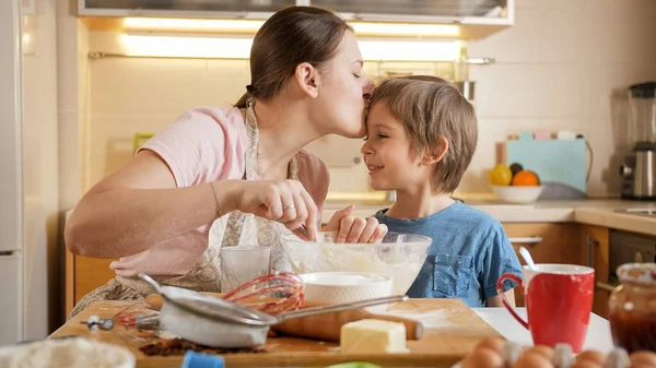Szczęśliwa matka całuje swojego uśmiechniętego syna podczas gotowania w kuchni — Zdjęcie stockowe