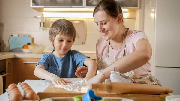 Madre sorridente con il figlioletto che spolvera la tavola di legno con la farina per fare rotolare la pasta dei biscotti. Bambini che cucinano con i genitori, piccolo chef, famiglia che ha tempo insieme, cucina domestica. — Foto Stock