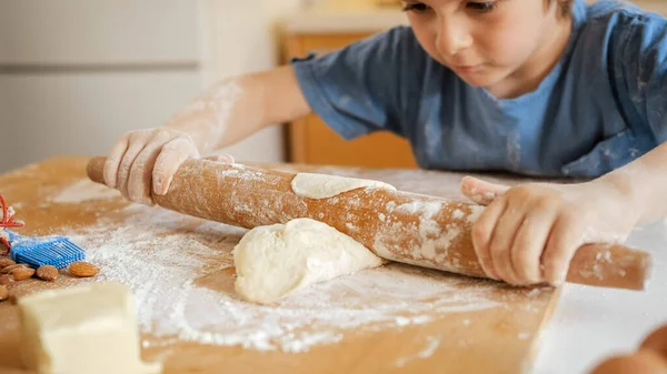 Маленький мальчик помогает матери и катит тесто на деревянной доске. Дети готовят с родителями, маленький шеф-повар, семья, проводящая время вместе, домашняя кухня. — стоковое фото