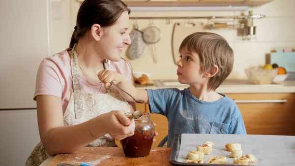 Ragazzino sorridente che nutre sua madre con marmellata di cucchiaio mentre prepara biscotti a casa. Bambini che cucinano con i genitori, piccolo chef, famiglia che ha tempo insieme, cucina domestica. — Foto Stock