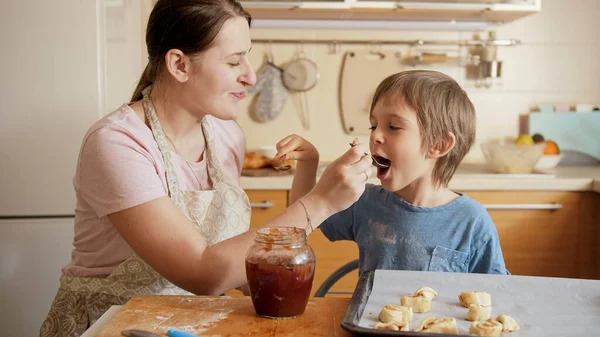 Felice madre sorridente con il piccolo figlio che mangia marmellata dolce dal barattolo mentre cuoce biscotti a casa. Bambini che cucinano con i genitori, piccolo chef, famiglia che ha tempo insieme, cucina domestica. — Foto Stock