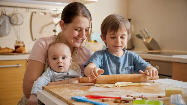 Młoda matka trzyma dziecko pomagając starszemu synowi piec pizzę w domu. Dzieci gotujące z rodzicami, mały kucharz, rodzina spędzająca razem czas, kuchnia domowa. — Zdjęcie stockowe