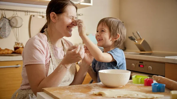料理をしながら小麦粉を投げながら母と遊ぶと幸せな笑顔の男の子。両親、小さなシェフ、家族と一緒に時間を過ごす子供たち、家庭用キッチン. — ストック写真