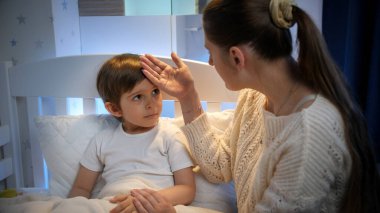 Genç bir anne, küçük oğluna dokunuyor ve onu alnından öpüyor. Çocuk hastalığı, hastalık ve ebeveyn bakımı kavramı