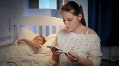 Diğer genç hasta oğlunun ateşini ölçüyor ve telefonla doktoru arıyor. Çocuk hastalığı, hastalık ve ebeveyn bakımı kavramı