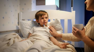 Bakıcı bir anne, hasta oğlunun ateşini ölçüyor ve ona sıcak çay veriyor. Çocuk hastalığı, hastalık kavramı. veli bakımı ve ev içi tedavi.