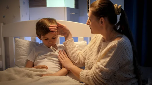 Opiekuńcza matka mierząca temperaturę swojemu choremu synowi leżącemu w łóżku w nocy. Pojęcie choroby dzieci, choroby i opieki rodzicielskiej. — Zdjęcie stockowe