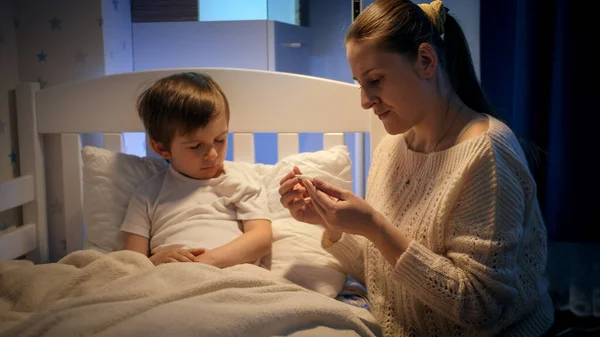 Young caring mother measuring temperature and kissing her sick little boy lying in bed. Concept of children illness, disease and parent care. — Stock Photo, Image