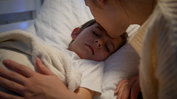 Menino bonito dormindo enquanto mãe carinhosa beijando-o e ajustando cobertor. Conceito de parentalidade e felicidade familiar. — Fotografia de Stock