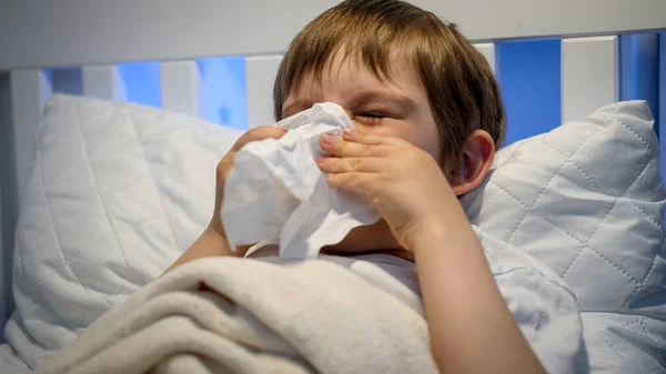 Retrato de um menino doente com corrimento nasal na cama e nariz a assoar no tecido de papel. Conceito de doença infantil, doença e cuidados de saúde. — Fotografia de Stock