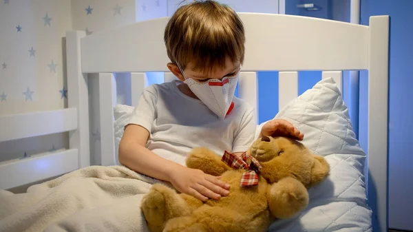 Liten pojke bär skyddande medicinsk mask liggande i sängen och leka med nalle. Begreppet barn virus och barn skydd under coronavirus Covid-19 pandemi. — Stockfoto