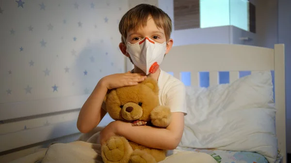 Menino bonito usando máscara médica protetora segurando e acariciando seu ursinho de pelúcia na cama à noite. Conceito de proteção contra vírus infantil e crianças durante a pandemia de coronavírus Covid-19. — Fotografia de Stock