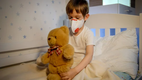 Menino sentindo-se doente usando máscara respiratória médica e brincando com ursinho de pelúcia. Conceito de proteção contra vírus infantil e crianças durante a pandemia de coronavírus Covid-19. — Fotografia de Stock