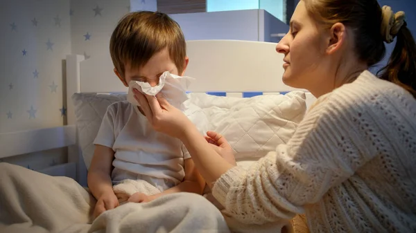 Egy beteg kisfiú, aki papírzsebkendőbe fújja az orrát, és lefekvés előtt beszél anyával. A gyermekbetegség, a betegség és a szülői ellátás fogalma — Stock Fotó