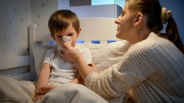 Madre premurosa soffia naso che cola del suo piccolo figlio malato sdraiato a letto. Concetto di malattia dei bambini, malattia e cura dei genitori. — Foto Stock