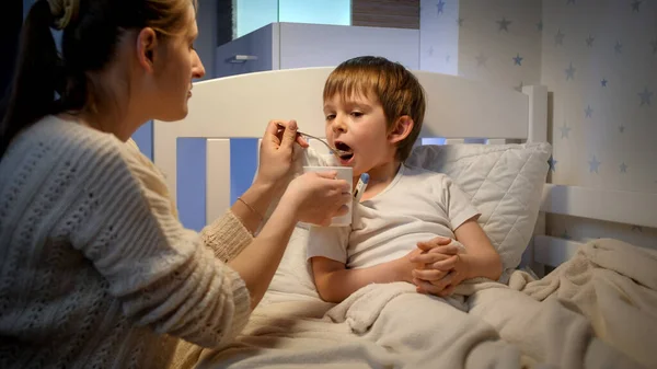 Η νεαρή μητέρα δίνει φάρμακα και σιρόπι από το κουτάλι στον άρρωστο γιο της ξαπλωμένο στο κρεβάτι. Έννοια της ασθένειας, της ασθένειας και της φροντίδας των γονέων — Φωτογραφία Αρχείου