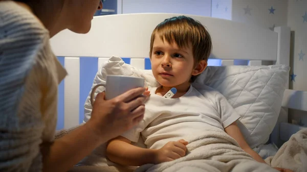 Mor ger varmt te till sin sjuka son mår dåligt liggande i sängen. Begreppet barnsjukdom, sjukdom. Föräldravård och hemvård. — Stockfoto