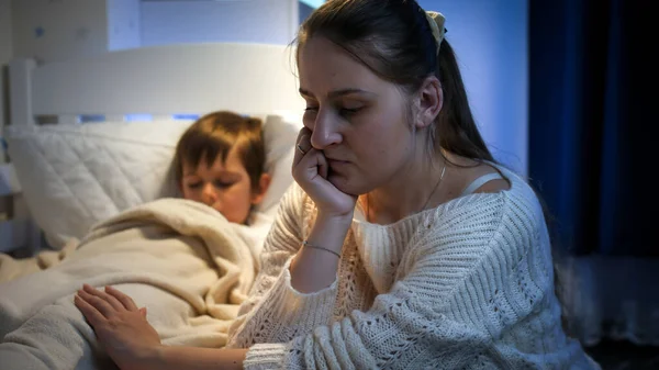 Madre molesta, triste y estresada sentada al lado de la cama de su hijo dormido por la noche. Concepto de madre solitaria, problemas familiares y depresión materna. —  Fotos de Stock