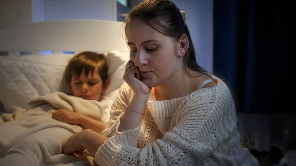 A fiatal szomorú nő aggódik a beteg fia miatt, aki az ágyban alszik. A magányos anya fogalma, családi problémák és anyai depresszió. — Stock Fotó