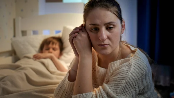Расстроенная и напряженная мать плачет рядом с кроватью своего маленького сына, спящего по ночам. Концепция одинокой матери, семейных проблем и материнской депрессии — стоковое фото