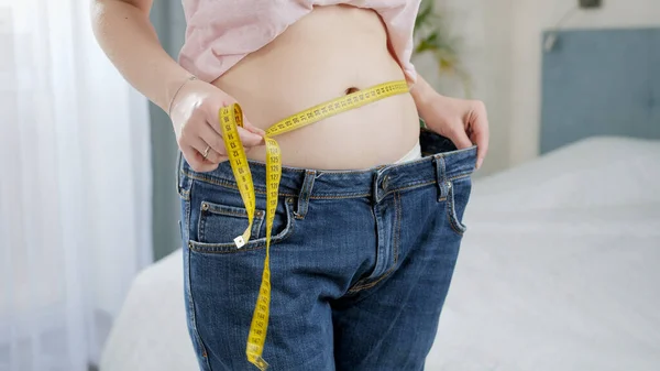 Piękna młoda kobieta z cienkim brzuchem mierzącym talię z taśmą i pokazująca duże dżinsy w lustrze. Pojęcie diety, utraty wagi i zdrowego stylu życia. — Zdjęcie stockowe