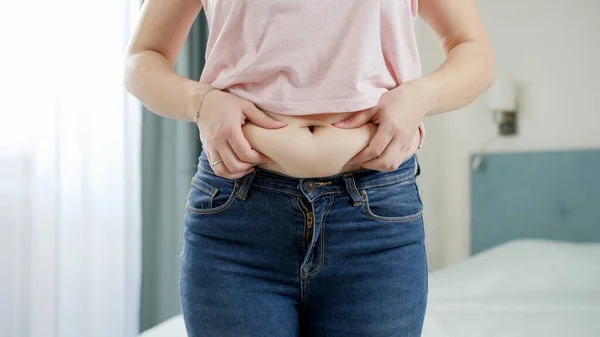 CLoseup of young woman in jeans holding fat fold on her belly. Concept of excessive weight, obese female, dieting and overweight problems — Stock Photo, Image