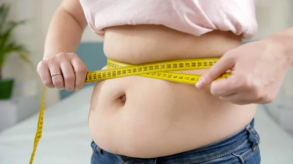 Крупный план толстой женщины, измеряющей свой большой живот желтой мерной лентой. Концепция питания, нездорового образа жизни, избыточного веса и ожирения — стоковое фото