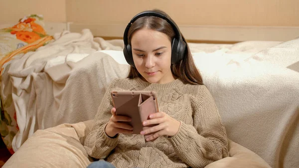Улыбающаяся девочка-подросток слушает музыку в наушниках с помощью планшетного компьютера в своей спальне — стоковое фото