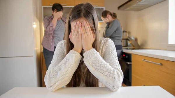 Расстроенная девушка плачет и закрывает лицо руками, потому что на ссоре и крики родителей на кухне. Семейное насилие, конфликты и проблемы в отношениях — стоковое фото