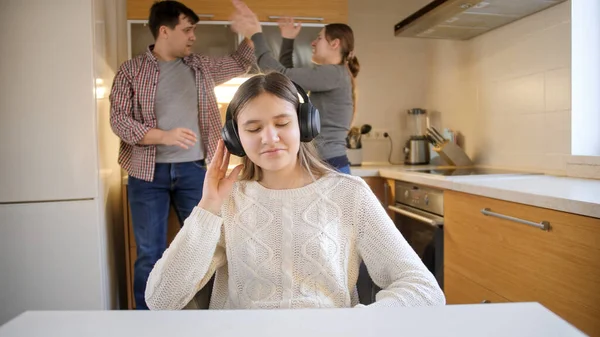 Teenage girl putting on headphones and enjoying music while parents shouting an arguing on the background. Family violence, conflicts and relationship problems — Stock Photo, Image