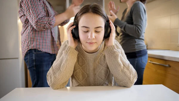 Adolescente fermant les oreilles avec des écouteurs pendant que les parents se disputent, crient et crient en arrière-plan. Violence familiale, conflits et problèmes relationnels — Photo