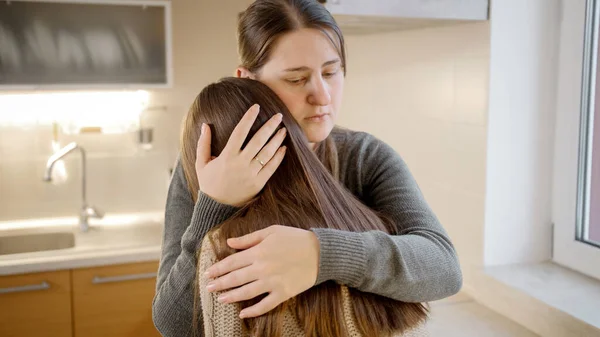 A fiatal anya ölelkezik és vigasztalja feldúlt síró lányát a konyhában. Szülői támogatás és vigasztaló gyermek — Stock Fotó