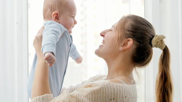 Portrait of young mother lifting up and smiling at her little baby son in front of big window at home. Concept of family happiness and child development. — Stock Photo, Image