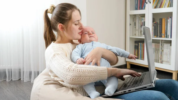 Fiatal anya simogatja a gyerekét, miközben az otthoni irodából dolgozik a laptopon. A családi boldogság és a gyermekfejlődés fogalma. — Stock Fotó