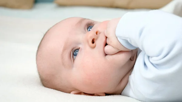 Retrato de niño adorable con ojos azules chupando su mano mientras está acostado en la cama — Foto de Stock