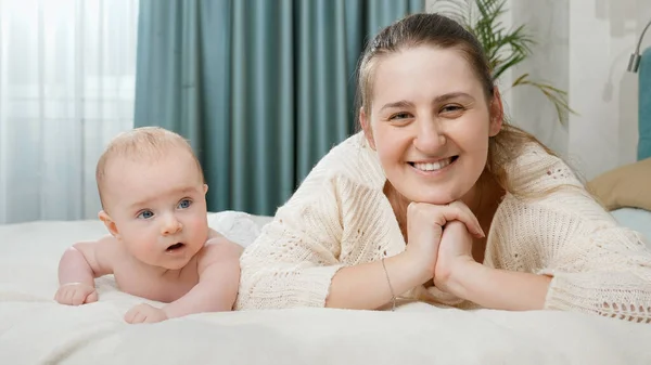 Young smiling happy mother lying with her baby in bed and looking in camera. Concept of parenting ,baby care and family happiness — Stock Photo, Image