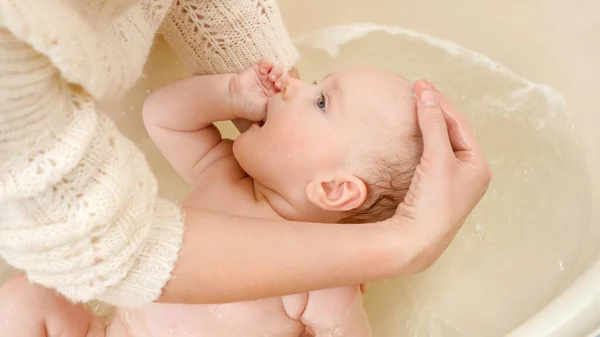 Frau hält und wäscht ihren kleinen Sohn in einer kleinen Plastikwanne. Konzept der Kindererziehung, Babypflege und Gesundheitsfürsorge. — Stockfoto