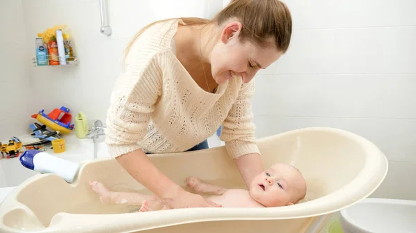 Feliz mamá sonriente lavando a su pequeño bebé en baño de plástico en casa — Foto de Stock