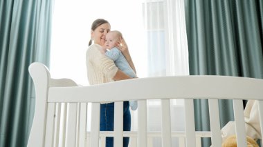 Gülümseyen bebek ve anne, sabah büyük bir pencereyle yatak odasında kucaklaşıyorlar. Ebeveynlik kavramı, aile mutluluğu ve bebek gelişimi