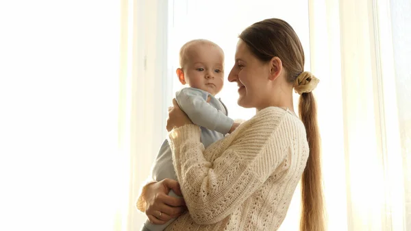 Portrait of loving young mother smiling at her little baby son against big window on sunset. Concept of family happiness and parenting — Stock Photo, Image