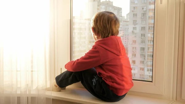 Liten pojke sitter på fönsterbrädan och tittar på gatan och solnedgången himlen genom fönstret. — Stockfoto