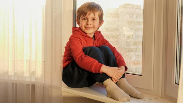 Retrato de un niño sonriente sentado en el alféizar de la ventana y mirando a la cámara. COncept de quedarse en casa y covid-19 pandemia — Foto de Stock