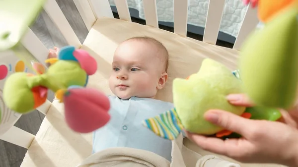 Молодая мать показывает красочные плюшевые игрушки своему маленькому сыну, лежащему в кроватке. Концепция детского образования, здравоохранения и развития — стоковое фото