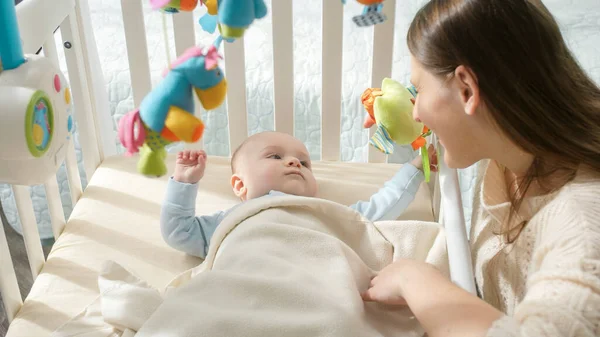 Happy smiling mother sitting at cradle and looking on her little baby son. Concept of parenting, family happiness and baby development — Stock Photo, Image