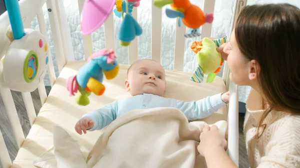 Lindo niño pequeño en la cuna mirando a la madre y coloridos juguetes de peluche. Concepto de paternidad, felicidad familiar y desarrollo del bebé — Foto de Stock