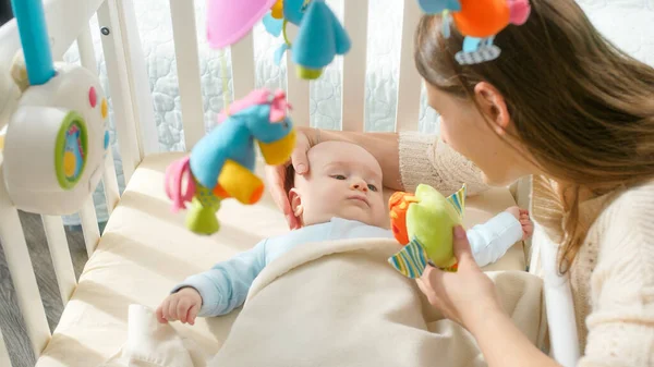 Feliz madre sonriente mostrando juguetes a su pequeño hijo acostado en la cuna. Concepto de paternidad, felicidad familiar y desarrollo del bebé — Foto de Stock